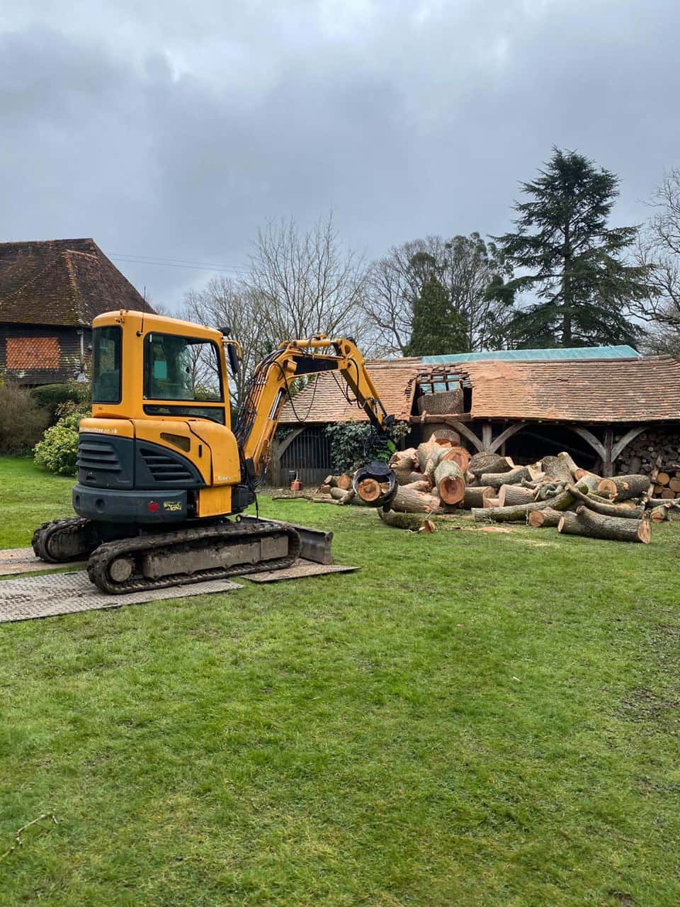 This is a photo of a tree which has grown through the roof of a barn that is being cut down and removed. There is a digger that is removing sections of the tree as well. Cotgrave Tree Surgeons